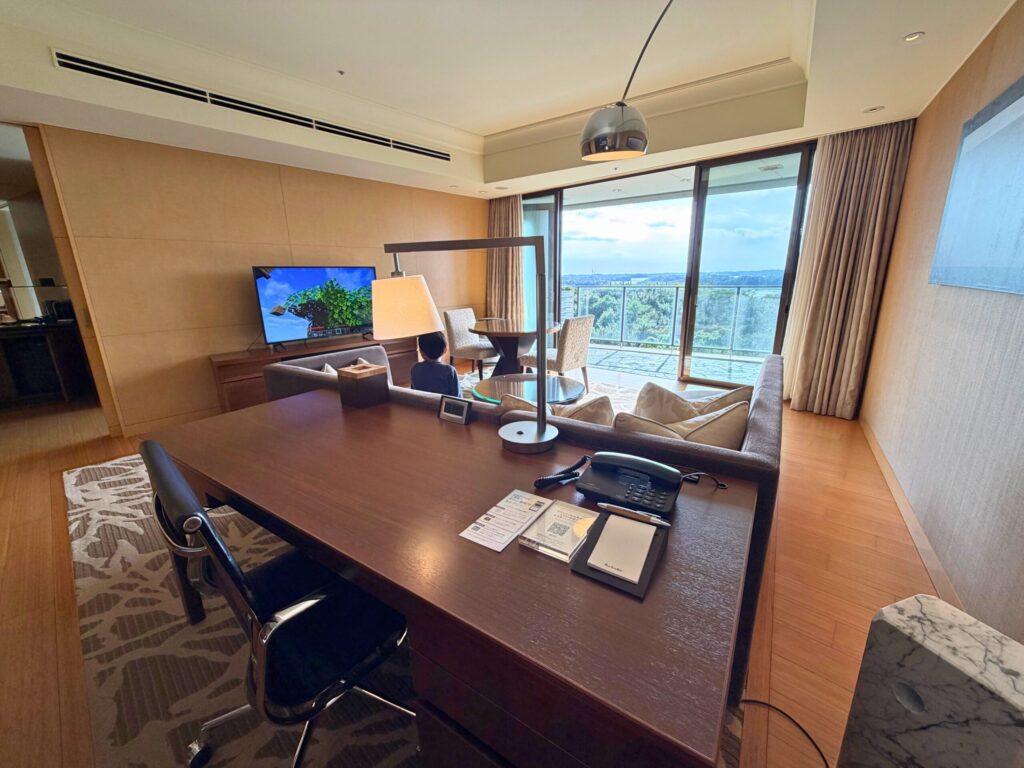 志摩観光ホテル ザ・ベイスイートのリビングルーム。英虞湾ビューと広いデスクがあり、子どもがテレビを楽しむファミリー滞在の様子。