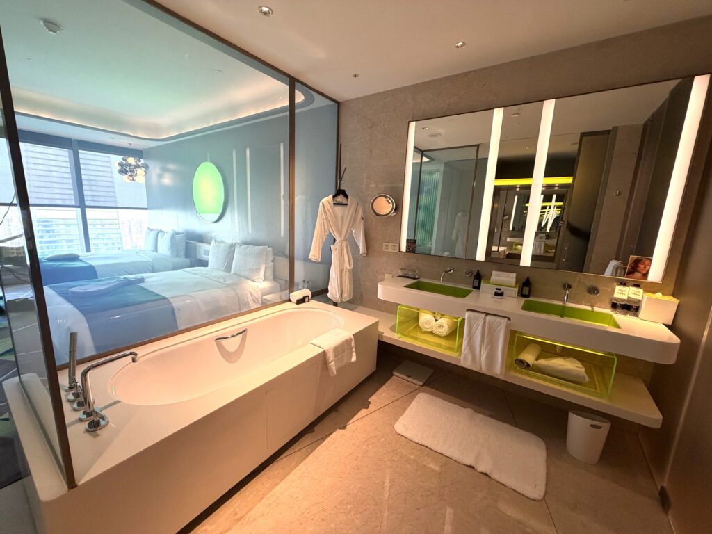 W蘇州ホテルの客室バスルーム。大きな浴槽とガラス壁越しに見えるベッドルーム、黄緑色のLEDアクセントライトが映えるダブルシンク