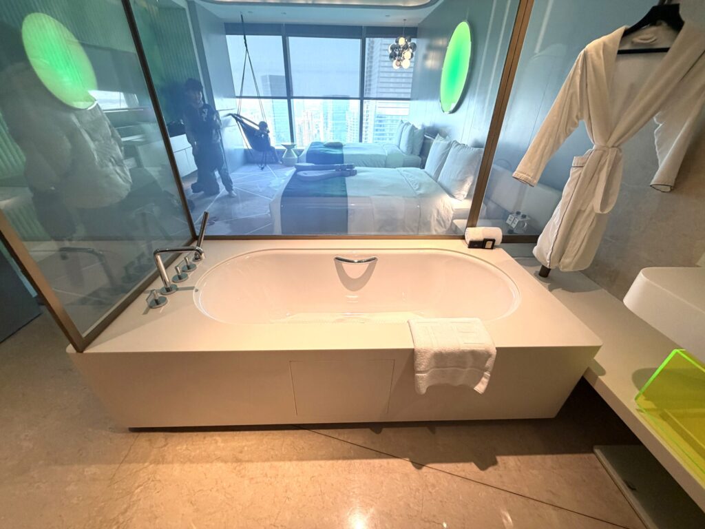 W蘇州ホテルのソーキングタブ。ガラス越しに緑の丸型ライトと蘇州の都市景観が広がり、白いバスローブがハンガーに掛かる