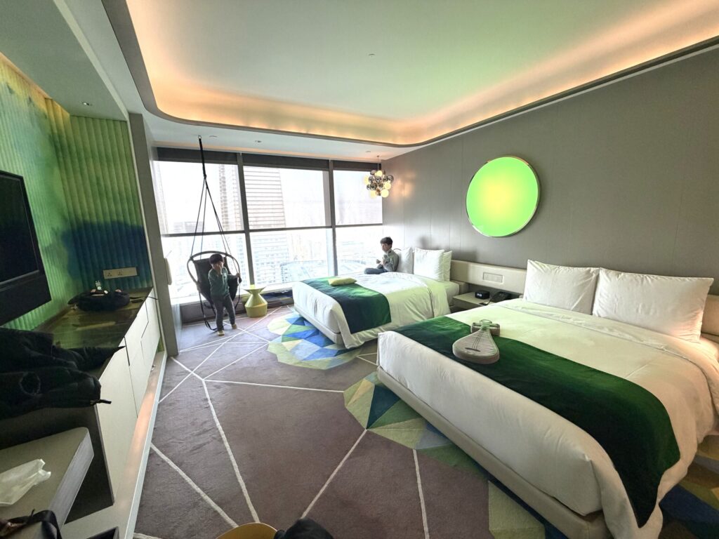 W蘇州ホテルのツインルーム。子どもがベッドに座り、緑のアクセントカラーと大きな窓から蘇州の高層ビル群を望む家族向け客室